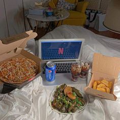 a table topped with pizza and other foods next to a laptop on top of a bed