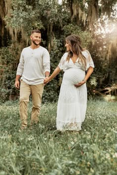 a pregnant couple holding hands and walking through the grass in front of trees with spanish moss