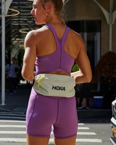 a woman in purple shorts is holding her waist bag and looking at the camera while waiting for someone to cross the street