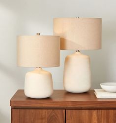 two white lamps sitting on top of a wooden table next to a bowl and lamp shade