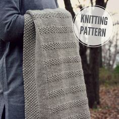 a woman holding up a knitted scarf in front of trees with leaves on the ground