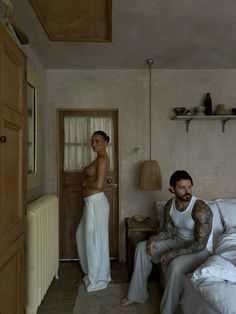 a man and woman sitting on a bed in a room with white linens, looking at each other