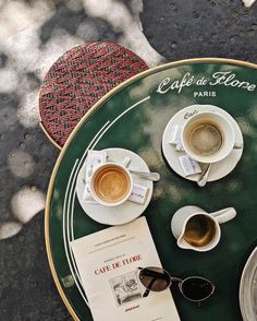 two cups of coffee are sitting on a table next to an open book and sunglasses