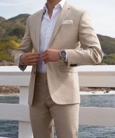 a man in a tan suit and white shirt is standing on a bridge by the water