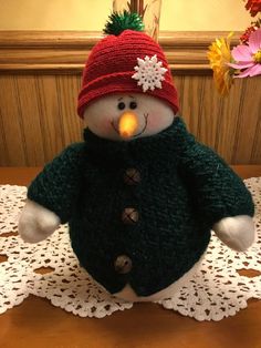 a stuffed snowman with a red hat and green sweater on top of a doily