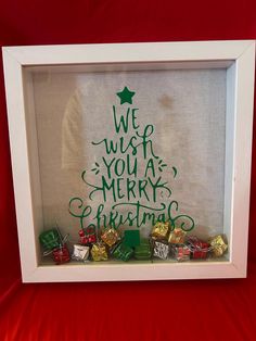 a christmas tree is framed in a white shadow box with green writing on it and wrapped presents underneath