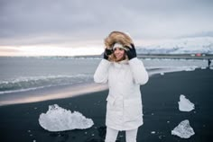 a woman standing on the beach wearing a white coat and holding a fur hat over her head