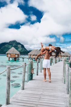 a man and woman are standing on a dock in front of some water with thatched huts