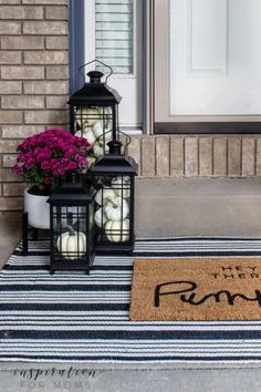 two lanterns are sitting on the front porch with flowers and pumpkins in them next to a welcome mat