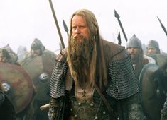 a man with long hair and beard standing in front of other men wearing armor holding swords