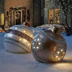 two christmas baubles sitting in the snow next to a house decorated with lights