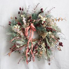 a wreath with dried flowers and greenery hanging on a white sheeted wall behind it