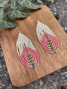 the pink and white beaded earrings are on a wooden board next to a plant