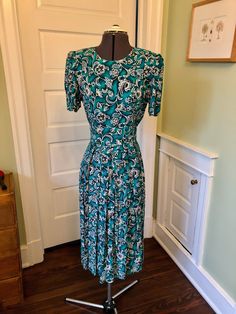 This day dress gives you a nice hourglass shape! This dress is made of 100% rayon floral fabric in emerald green, blue and ivory. It features a crew neckline, short sleeves, a front waist panel with covered buttons, back waist is elasticized, pleated skirt, and back zipper closure. It is unlined. There are shoulder pads that extend into the sleeves, creating a pleated drop. This garment is clean, with no stains or snags. Measurements to guide you: Bust: 36" Waist: 25-30" Hip: 50+" Sleeve: 10.5" Length from top of shoulder to hem: 46" Decade: 80s Fabric: Rayon Size: M Condition: Great Brand: Karin Stevens Petites It's a delight to pick through time for you to find these vintage treasures. Green A-line Midi Dress With Floral Print, Green A-line Floral Dress For Garden Party, Fitted Dark Green Summer Dress, Elegant Green Floral Short Sleeve Dress, Elegant Short Sleeve Green Floral Dress, Elegant Green Floral Dress With Short Sleeves, Elegant Green Short Sleeve Floral Dress, Green Floral Knee-length Dress For Garden Party, Green Knee-length Floral Dress For Garden Party