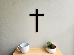 a wooden table topped with two white cups and a cross on the wall behind it