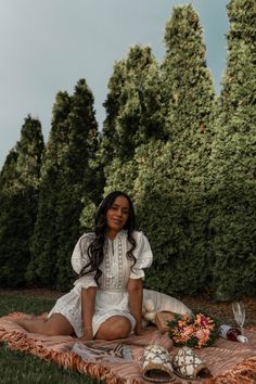 a woman sitting on top of a blanket in the middle of a field next to trees