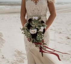 a woman standing on top of a beach holding a bouquet of flowers and greenery