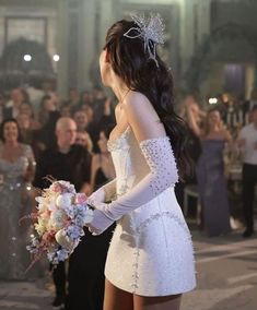 a woman in a short white dress holding a bridal bouquet and looking at the crowd