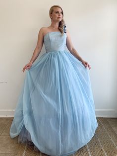 "This is a vintage 1950's light blue tone wedding dress. It is gives strong Cinderella vibes with the color and fit. The dress has a synthetic light blue tone lining, a layer of blue tone tulle, and a soft chiffon-type outer layer in the same light blue tone. The bodice is fitted and has a side zipper. The skirt has small pickups at the hem accentuated by a medium blue tone velvet bow. The dress also has a one shoulder strap finished in a bow in the same medium blue tone velvet. The dress has a Light Blue Ball Gown With Fitted Bodice For Wedding, Vintage Blue Dress For Wedding, 1950s Style Blue Wedding Dress, Blue Tone Wedding, Small Pickups, Blue Tone, Vintage Gowns, Wrap Shawl, Velvet Bow