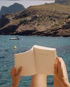 a person is reading a book while sitting on a boat in the water with mountains behind them