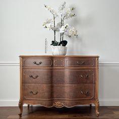 a vase with flowers on top of a wooden dresser next to a white wall and hardwood floor