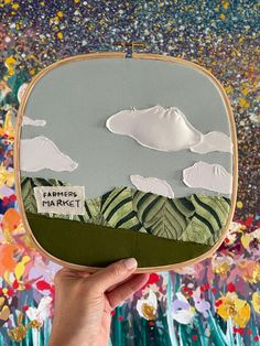 someone is holding up an embroidered artwork with clouds and mountains in the sky above them