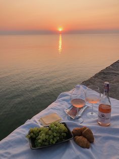 two glasses of wine and some grapes on a table by the water at sunset or sunrise