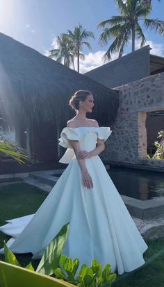 a woman standing in front of a pool wearing a wedding dress with an off the shoulder neckline