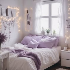 a white bed with purple pillows and blankets in a room decorated with fairy lights on the wall