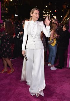 a woman in white pants and jacket waves to the crowd on pink carpet at an event