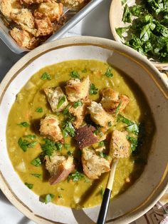 a white bowl filled with soup next to some bread and greens on a table top