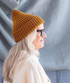 a woman wearing a knitted hat and glasses looks off into the distance while standing in front of a curtain