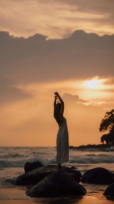 a woman standing on rocks in front of the ocean with her arms raised up to the sky