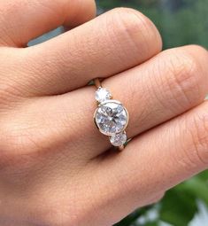 a woman's hand with a diamond ring on top of her finger and green plants in the background