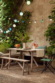 an outdoor table and bench with lights strung from the ceiling above it, surrounded by greenery