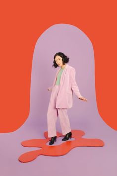 a woman standing in front of a purple and red background wearing a pink suit with black heels