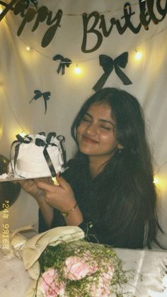 a woman sitting at a table with a cake in front of her and lights on the wall behind her