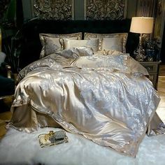 a bed with silver sheets and pillows in a room next to a lamp on a table