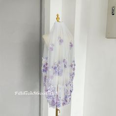 a white veil with purple flowers hanging from it's side on a stand in front of a wall