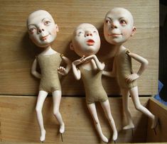 three small dolls are posed in front of a wooden surface, one is wearing a tan bodysuit and the other has white skin