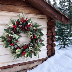 a wreath is hanging on the side of a cabin in the snow with evergreens and red berries