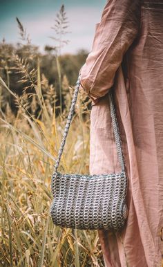 a woman is standing in the tall grass holding a woven handbag on her shoulder