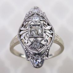 An old European cut diamond is centered on this antique 14k white gold filigree ring. The 3mm .10 carat precious gem is mounted in a square setting. Delicate airy scroll filigree with leaves and millgrain dots decorates the ring. The 13/16 inch long face has a domed gallery with engraved profiles, which is emulated on the ring's shoulders. There are four additional 1.5mm to 1.8mm diamonds set around on the ring. Collectively, this ring has an estimated total weight of .18 cwt diamonds.  The insi Antique Platinum Diamond Ring With Diamond Accents, Antique Diamond White Diamond Ring, Victorian Diamond Ring In White Gold, Victorian White Gold Diamond Ring, Victorian Style White Diamond Ring, Victorian Diamond Ring With Diamond Accents, Victorian Diamond White Diamond Ring, Victorian Style Diamond Ring With Accents, Victorian Style Diamond White Diamond Ring