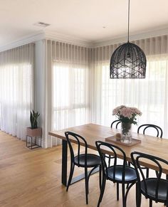 a dining room table with four chairs and a vase on top of it, in front of sheer curtains