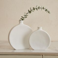 two white vases sitting on top of a table next to each other with green leaves in them