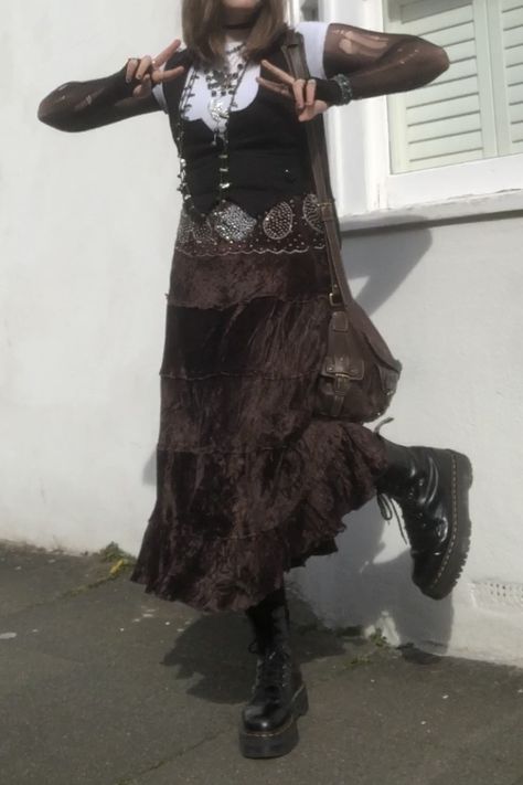 A mid-size person/girl wearing an oversized white fairy crop top, a black waistcoat over it, a black ripped tights top under it, a long dark brown skirt, ripped rose-patterned tights under it, black platform dr. Martens and multiple crystal rings and bracelets. 	The person is also wearing multiple silver necklaces; one in form of a fairy and one in form of a moon, a brown rosary-style and a green bead necklace and a black lace choker. They also have a brown handbag with them. Brown Goth Aesthetic, Brown Goth Outfit, Maxi Skirt Outfit Grunge, Dark Academia Goth Outfit, Crow Core Aesthetic Outfits, Crow Core Outfit, Cottage Goth Fashion, Steampunk Aesthetic Outfit, Dark Cottage Core Outfits