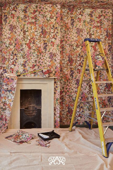 Room being decorated with floral pink wallpaper. There is a yellow ladder, dust sheets, a roller on the floor and a white fireplace. Colorful Home Wallpaper, Wallpaper For Built Ins, Wallpaper That Goes With Everything, Big Pattern Wallpaper, Rooms With Wallpaper Ideas, Hacienda Wallpaper, Partial Wallpaper, Fun Wallpaper Accent Wall, Painterly Wallpaper