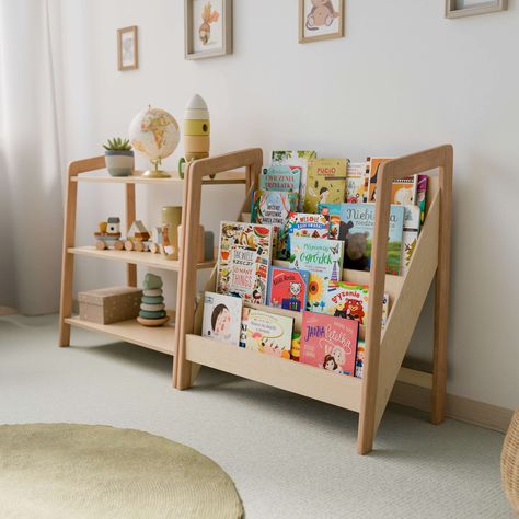 The image shows a Montessori-style setup featuring a bookshelf and a toy storage shelf, both made of natural wood. The bookshelf has multiple tiers displaying a variety of colorful children's books with their covers facing outward for easy selection. The toy storage shelf has three tiers, displaying items such as a globe, a rocket-shaped stacking toy, wooden vehicles, and decorative containers. Book Storage For Children, Bookcase Nursery Ideas, Children Room Furniture, Baby Room Toy Storage Ideas, Montessori Toy Organization, Children Book Storage, Toddler Bedroom Toy Storage, Montisorri Baby Room, Kids Toys Storage Ideas