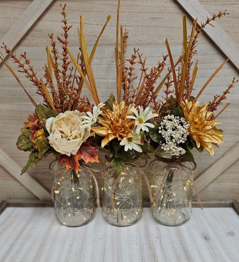 These beautiful 3 piece quart size mason jar centerpiece set includes three sets of warm white LED fairy lights, three jute bows, and three deluxe faux fall arrangements. These centerpieces are very popular for weddings, baby showers, parties, corporate events, holiday events, and any other event that requires table centerpieces. They also look great in any room of your house or office. To see other lighted jar options and all of my other listings, please view my shop: https://www.etsy.com/shop/GlassyGalDecor  Each jar set includes: (1) clear quart size mason jar (1) set of LED fairy lights (1) Silk fall flower arrangement (1) jute bow Dimensions:  Quart jars are approximately 6.25" tall (approximately 15" with flowers) **The LED fairy lights last for 24-48 continuous hours before needing Mason Jar Decorations For Weddings, Fall Birthday Centerpieces For Table, Fall Color Birthday Decorations, Centerpieces That Are Not Flowers, Tall Fall Centerpieces Wedding, Fall Color Party Decorations, Fall Baby Shower Centerpieces For Girl, Mason Jar Fall Centerpieces, Mason Jar Centerpieces For Weddings