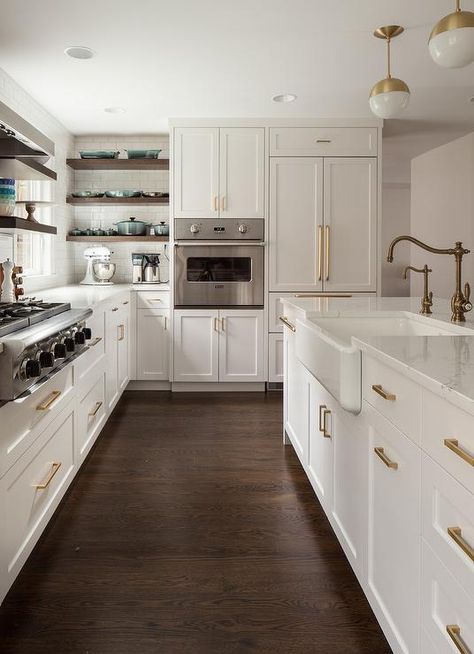 Gorgeous kitchen boasts white and gold pendants illuminating a white center island fitted with a farmhouse sink and antique brass hook and spout faucet. Kitchen With Brown Wood Floors, White Kitchen Dark Brown Floor, White Kitchen Cabinets With Brown Floor, Kitchen Brown Floor White Cabinets, White Kitchen With Dark Brown Floors, Small Kitchen Dark Floor, Dark Floors Kitchen Ideas, Dark Floors With Dark Cabinets, Kitchen Remodel Dark Wood Floors